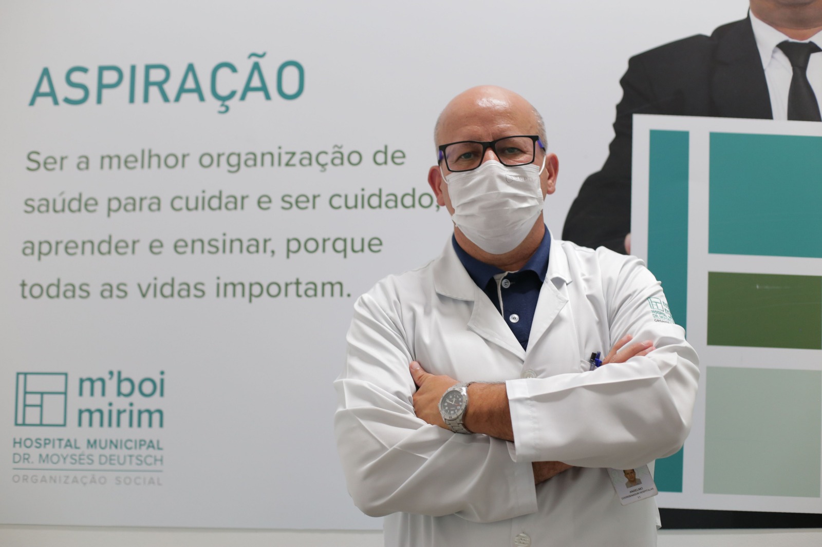 Na imagem, Anselmo está de braços cruzados, usando jaleco branco e óculos, e máscara de proteção. Ao fundo, um painel  com uma mensagem do Hospital Municipal M'Boi Mirim